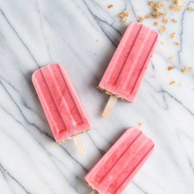 overhead image: 3 strawberry yogurt popsicles with granola on gray marble background
