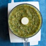 Salsa verde in a food processor on a blue backdrop.