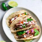 Three grilled flank steak tacos on an oval plate, surrounded by recipe ingredients.