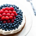 Bright photo of a white cake topped with a thick layer of cream cheese frosting, topped with raspberries in the center and blueberries toward the edge.