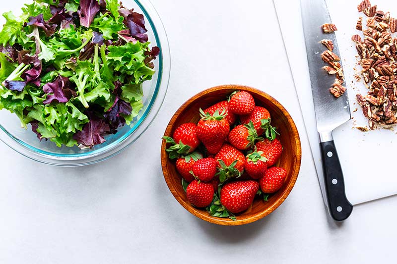 A bowl of strawberries next to a bowl of mixed greens and a cutting board with chopped pecans and a knife. 